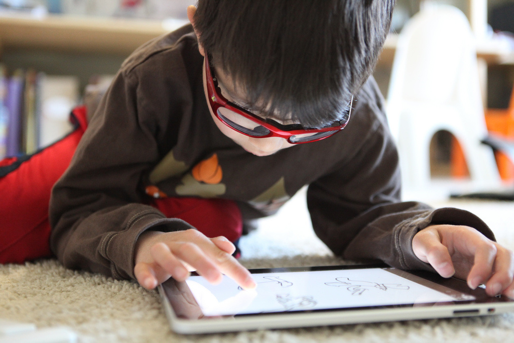 A young boy with glasses is laying on the floor interacting with an iPad/tablet.