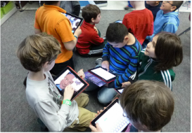 A group of eight boys are interacting while holding/using iPads and tablets.