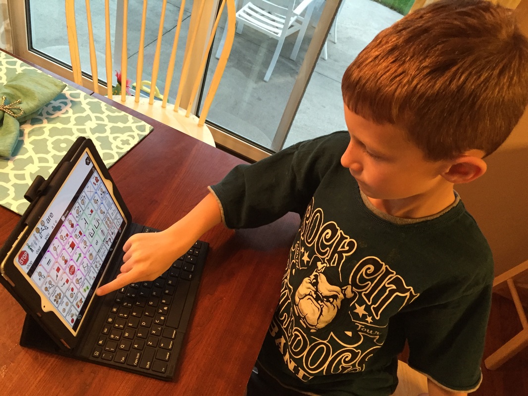 A young boy is sitting at a kitchen table interacting with a communication app on an iPad.